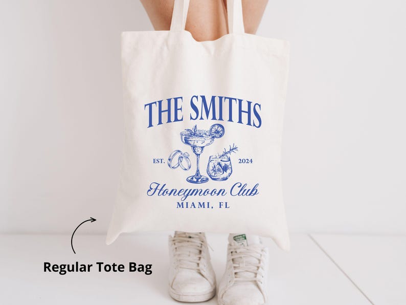 May include: Cream-colored tote bag with blue text and illustrations. The text reads "THE SMITHS Honeymoon Club Miami, FL" with the year 2024. Illustrations include a cocktail, rings, and a drink. The bag has two handles and is held up.