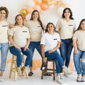 K&ouml;nnte beinhalten: Sieben Frauen tragen cremefarbene T-Shirts mit schwarzem Text, darunter "Bride", "Destiny", "Sarah", "Emily", "Leanna" und "Loretta". Sie stehen vor einem Hintergrund aus Pfirsich- und Goldballons.