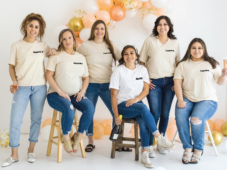 K&ouml;nnte beinhalten: Sieben Frauen tragen cremefarbene T-Shirts mit schwarzem Text, darunter "Bride", "Destiny", "Sarah", "Emily", "Leanna" und "Loretta". Sie stehen vor einem Hintergrund aus Pfirsich- und Goldballons.