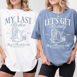 May include: Two white t-shirts with blue text and graphics. The first shirt says "My Last Rodeo" with a boot and a drink graphic. The second shirt says "Let's Get Rowdy" with a boot and a drink graphic. Both shirts say "Amy's Bachelorette Nashville, TN".