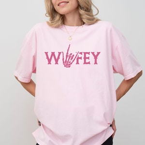 May include: A light pink t-shirt with the word "WIFEY" in a dark pink, gothic-style font. The letter "F" is replaced with a skeleton hand giving the middle finger. The shirt is a casual, oversized fit.