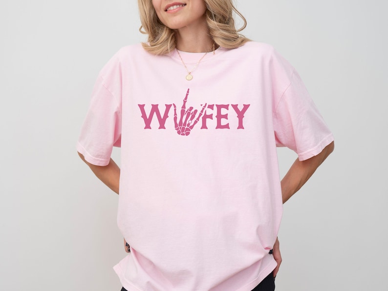May include: A light pink t-shirt with the word "WIFEY" in a dark pink, gothic-style font. The letter "F" is replaced with a skeleton hand giving the middle finger. The shirt is a casual, oversized fit.