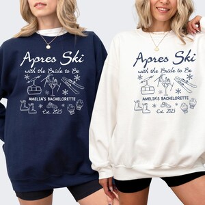 Könnte beinhalten: Marineblaue und cremefarbene Sweatshirts mit dem Text "Apres Ski with the Bride to Be" und "Amelia's Bachelorette Est. 2025". Das Design umfasst Skilift-, Ski- und Schneeflocken-Grafiken. Die Sweatshirts sind ein bequemer, lässiger Stil.
