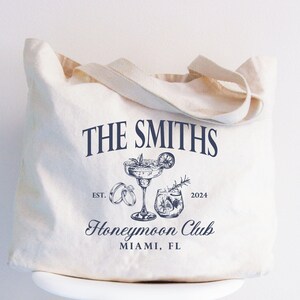 May include: Cream-colored tote bag with the text "THE SMITHS" in navy blue. Below the text is a graphic of a cocktail, rings, and a drink. The words "Honeymoon Club" and "MIAMI, FL" are also printed on the bag.
