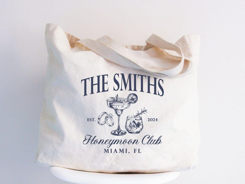 May include: Cream-colored tote bag with the text "THE SMITHS" in navy blue. Below the text is a graphic of a cocktail, rings, and a drink. The words "Honeymoon Club" and "MIAMI, FL" are also printed on the bag.