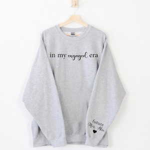 Puede incluir: Una sudadera gris claro con la frase "in my engaged era" impresa en negro en el pecho. La manga tiene el texto "future Mrs. Alex" con un peque&ntilde;o coraz&oacute;n negro. La sudadera est&aacute; colgada en una percha de madera.