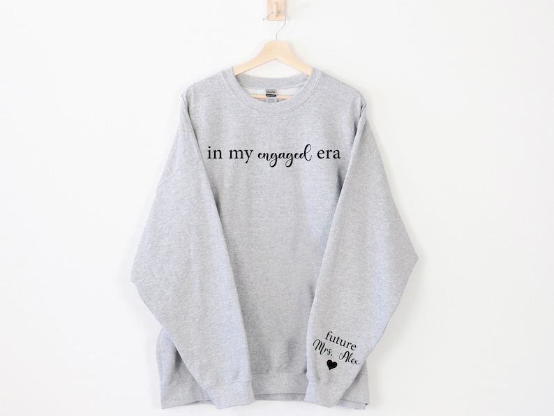 Puede incluir: Una sudadera gris claro con la frase "in my engaged era" impresa en negro en el pecho. La manga tiene el texto "future Mrs. Alex" con un peque&ntilde;o coraz&oacute;n negro. La sudadera est&aacute; colgada en una percha de madera.