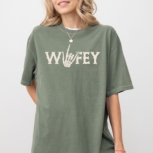 May include: Olive green t-shirt with the word "WIFEY" in a vintage font, featuring a skeleton hand making a gesture. The shirt has a relaxed fit and short sleeves. A gold necklace is visible.