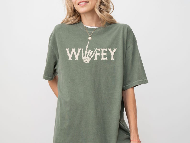 May include: Olive green t-shirt with the word "WIFEY" in a vintage font, featuring a skeleton hand making a gesture. The shirt has a relaxed fit and short sleeves. A gold necklace is visible.