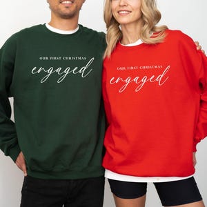 May include: Two crewneck sweatshirts, one in dark green and the other in red. Both have the text "OUR FIRST CHRISTMAS" above the word "engaged" in a cursive font. The sweatshirts are a festive choice for couples.