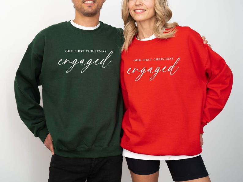 May include: Two crewneck sweatshirts, one in dark green and the other in red. Both have the text "OUR FIRST CHRISTMAS" above the word "engaged" in a cursive font. The sweatshirts are a festive choice for couples.
