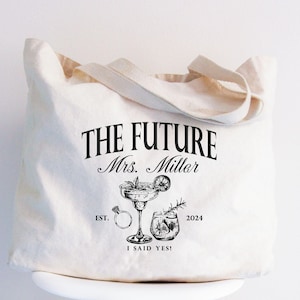 K&ouml;nnte beinhalten: Cremefarbene Tragetasche mit dem Text "THE FUTURE Mrs. Miller" und Illustrationen eines Cocktails, eines Rings und eines Getr&auml;nks. Der Text "I SAID YES!" und das Jahr 2024 sind ebenfalls auf der Tasche aufgedruckt. Die Tasche hat cremefarbene Griffe.
