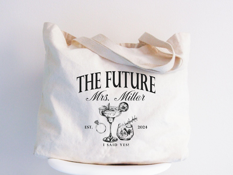 K&ouml;nnte beinhalten: Cremefarbene Tragetasche mit dem Text "THE FUTURE Mrs. Miller" und Illustrationen eines Cocktails, eines Rings und eines Getr&auml;nks. Der Text "I SAID YES!" und das Jahr 2024 sind ebenfalls auf der Tasche aufgedruckt. Die Tasche hat cremefarbene Griffe.