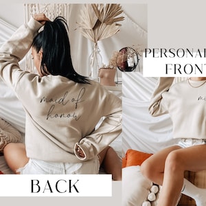 May include: A beige sweatshirt with the text "maid of honor" on the back and the text "mia" on the front. The sweatshirt is being modeled by a person.