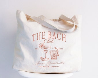 May include: Cream-colored tote bag with the text "THE BACH Club" in a reddish-brown font. Below the text are illustrations of two cocktails and the text "Amy's Bachelorette, Chandler, Arizona, 2021". The bag has matching handles.