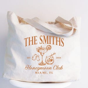 May include: Off-white canvas tote bag with brown text and illustrations. The text reads "THE SMITHS" above a cocktail glass, rings, and a drink. Below is "Honeymoon Club" and "MIAMI, FL". The year "2024" is also included.