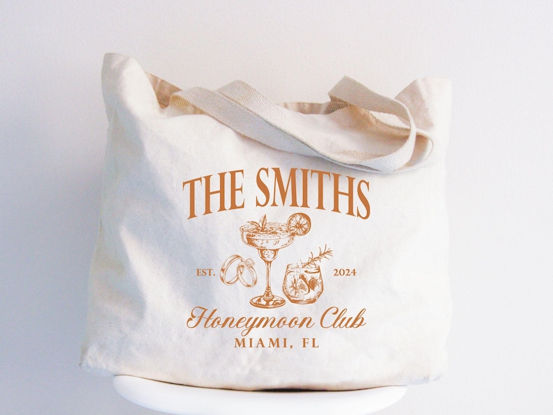 May include: Off-white canvas tote bag with brown text and illustrations. The text reads "THE SMITHS" above a cocktail glass, rings, and a drink. Below is "Honeymoon Club" and "MIAMI, FL". The year "2024" is also included.