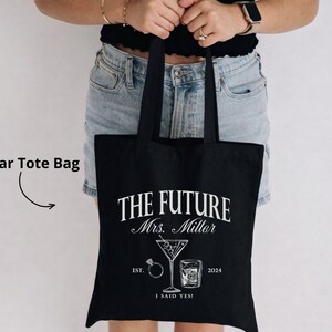K&ouml;nnte beinhalten: Schwarze Tragetasche mit wei&szlig;em Text und Grafiken. Der Text lautet "THE FUTURE Mrs. Miller" mit einem Martiniglas, einem Ring und einem Whiskeyglas. Darunter steht der Text "EST. 2024 I SAID YES!"