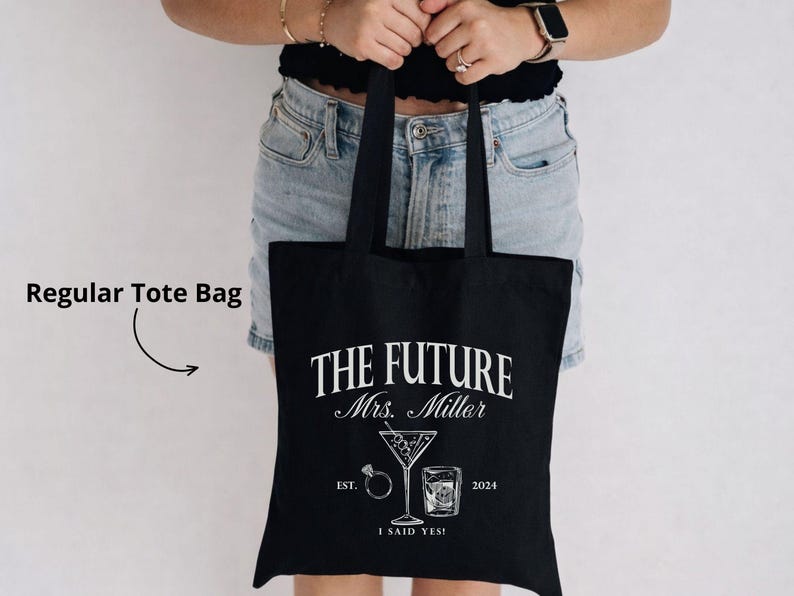 K&ouml;nnte beinhalten: Schwarze Tragetasche mit wei&szlig;em Text und Grafiken. Der Text lautet "THE FUTURE Mrs. Miller" mit einem Martiniglas, einem Ring und einem Whiskeyglas. Darunter steht der Text "EST. 2024 I SAID YES!"