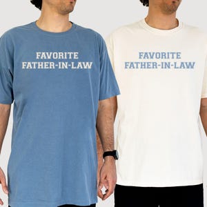 May include: Two short-sleeved t-shirts, one blue and one cream, each with the text "FAVORITE FATHER-IN-LAW" in light blue. The shirts are worn by individuals, suggesting casual wear. The text is centered on the chest.