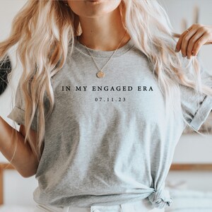 May include: A gray t-shirt with the text "In My Engaged Era 07.11.23" printed on the front.