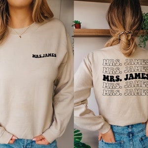 K&ouml;nnte beinhalten: Ein beiger Pullover mit dem Text "MRS. JAMES" auf der Vorder- und R&uuml;ckseite. Der Text auf der R&uuml;ckseite wird f&uuml;nfmal in einer Retro-Schrift wiederholt.