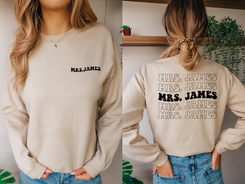 K&ouml;nnte beinhalten: Ein beiger Pullover mit dem Text "MRS. JAMES" auf der Vorder- und R&uuml;ckseite. Der Text auf der R&uuml;ckseite wird f&uuml;nfmal in einer Retro-Schrift wiederholt.