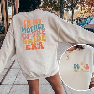 May include: A cream-colored sweatshirt with the text "In My Mother of the Bride Era" printed on the back in rainbow colors. The front of the sweatshirt has the text "MOB" in rainbow colors with the text "Mother of the Bride" underneath.