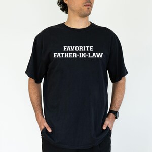 May include: Black t-shirt with the text "FAVORITE FATHER-IN-LAW" in white block letters. The shirt is a crew neck style and is being worn by a person with their hands in their pockets.