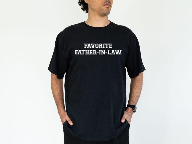 May include: Black t-shirt with the text "FAVORITE FATHER-IN-LAW" in white block letters. The shirt is a crew neck style and is being worn by a person with their hands in their pockets.