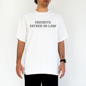 May include: White t-shirt with the text "FAVORITE FATHER-IN-LAW" in gray block letters. The shirt is a short-sleeved, crew neck style. The person is wearing a black watch and black pants.