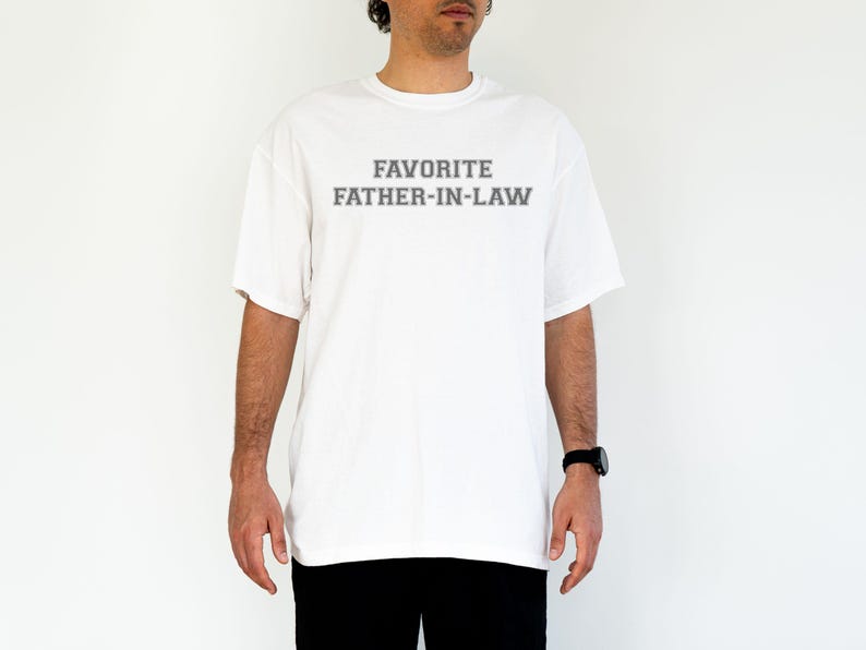 May include: White t-shirt with the text "FAVORITE FATHER-IN-LAW" in gray block letters. The shirt is a short-sleeved, crew neck style. The person is wearing a black watch and black pants.