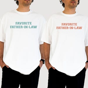 May include: Two white t-shirts with the text "FAVORITE FATHER-IN-LAW" printed on the front. One shirt has the text in teal, the other in coral. The models are wearing black pants and a black watch.