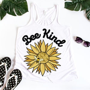 Könnte beinhalten: Weißes Tanktop mit einem Sonnenblumen-Motiv und dem Schriftzug "Bee Kind" in schwarzer Schrift. Die Sonnenblume hat gelbe Blütenblätter und eine Wabe mit einer kleinen Biene. Schwarz-weiß gepunktete Flip-Flops und Sonnenbrillen sind ebenfalls im Bild.