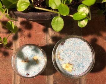 May include: Two glass jars filled with blue bath salts and dried orange peel. The jars are on a brick surface.