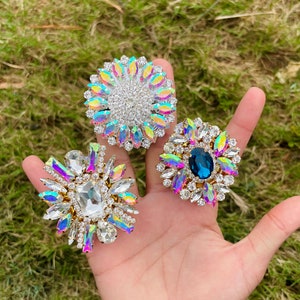 Könnte beinhalten: Drei schimmernde Strasssteine mit goldenen Fassungen. Die Verzierungen haben die Form einer Blume, einer Sternform und eines Kreises. Die Blume und die Sternform haben klare Strasssteine, während der Kreis einen großen blauen Strassstein in der Mitte hat.