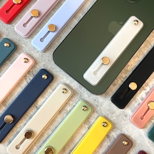 May include: An assortment of colorful phone grip stands in various shades, including red, lavender, blue, white, black, pink, green, yellow, brown, and burgundy. Each grip features a gold-colored circular button and a rectangular base.