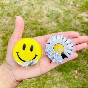 Puede incluir: Dos agarres para teléfono de plástico transparente con purpurina en el interior. Un agarre tiene una carita sonriente amarilla y el otro una margarita blanca.