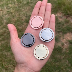May include: Four phone ring holders in different colors: rose gold, silver, black, and gold. The holders are on a hand.