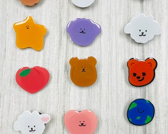 May include: A collection of colorful, cartoon-style charms in various shapes, including a planet, a peach, a cloud, and a milk carton. Each charm features a unique, smiling face design. The charms are arranged on a light gray, wood-grain surface.