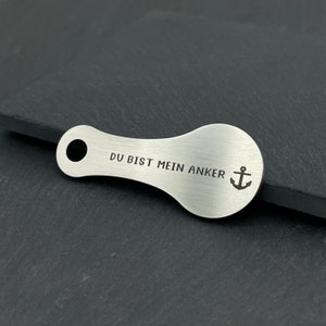 May include: A silver key chain with a teardrop shape and a hole at the top. The keychain has the text "DU BIST MEIN ANKER" and an anchor symbol engraved on it.