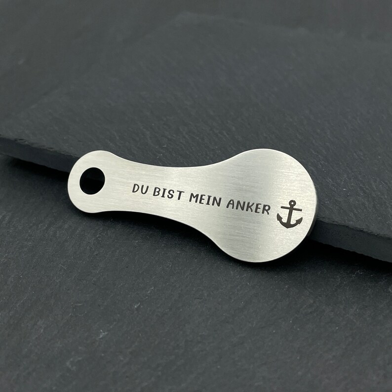 May include: A silver key chain with a teardrop shape and a hole at the top. The keychain has the text "DU BIST MEIN ANKER" and an anchor symbol engraved on it.