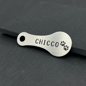 May include: Silver dog tag with the name "CHICCO" engraved on it and two paw prints.
