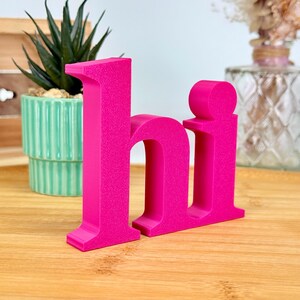 Könnte beinhalten: Hellrosa 3D-gedruckte Buchstaben, die das Wort "hi" bilden. Die Buchstaben stehen auf einer Holzoberfläche, mit einer Sukkulente in einem türkisfarbenen Topf und einer dekorativen Vase im Hintergrund. Die Buchstaben sind der Fokus.
