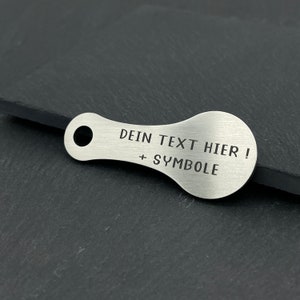 May include: A silver metal keychain with a rounded shape and a hole for attaching keys. The keychain has the text "DEIN TEXT HIER! + SYMBOLE" engraved on it.