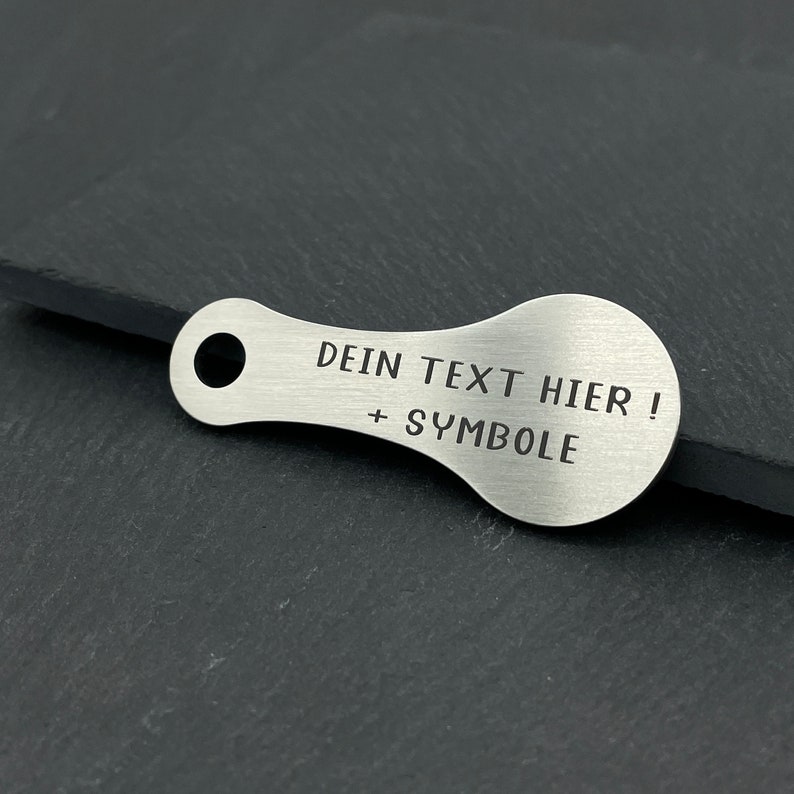 May include: A silver metal keychain with a rounded shape and a hole for attaching keys. The keychain has the text "DEIN TEXT HIER! + SYMBOLE" engraved on it.
