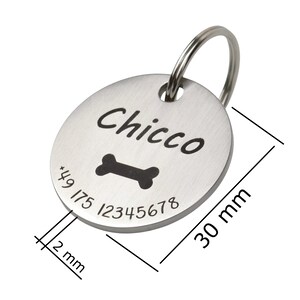 May include: A circular silver-toned metal pet ID tag featuring the engraved name Chicco, a bone icon, and the phone number +49 175 12345678. The tag includes a metal split ring and has visible dimensions of 30 mm in diameter and 2 mm in thickness.