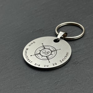 Peut inclure: Un porte-cl&eacute;s en argent avec un design de boussole et le texte "Dein Wunschtext bis zu 30 Zeichen".