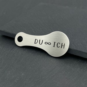 May include: A silver metal keychain with a rounded shape and a hole for attaching keys. The keychain is engraved with the text "DU &infin; ICH".