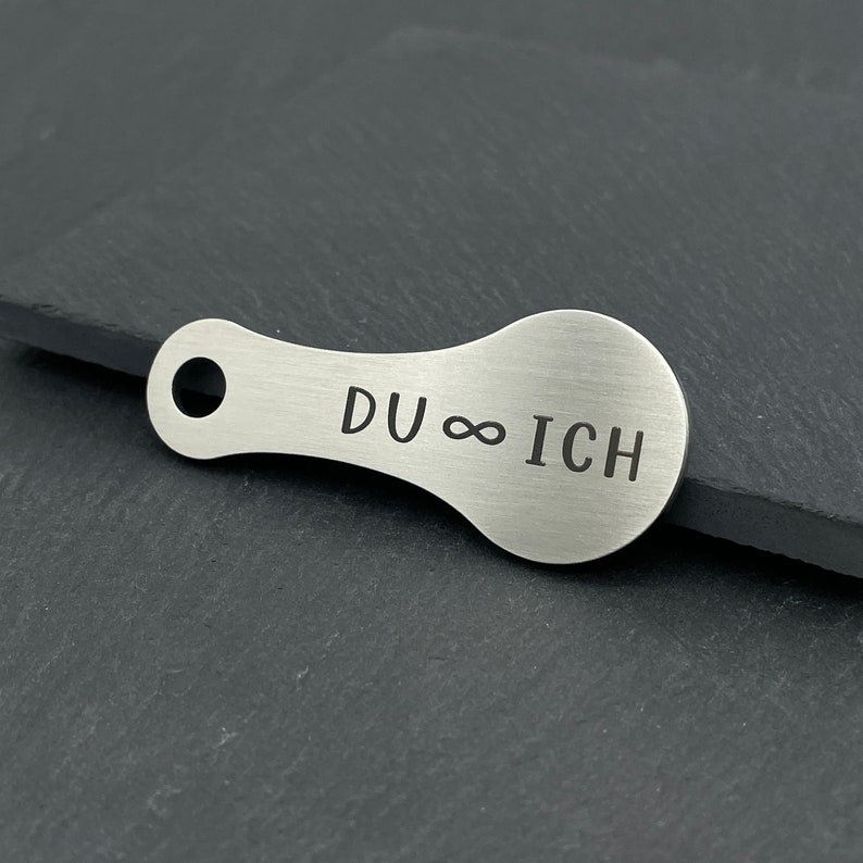 May include: A silver metal keychain with a rounded shape and a hole for attaching keys. The keychain is engraved with the text "DU &infin; ICH".